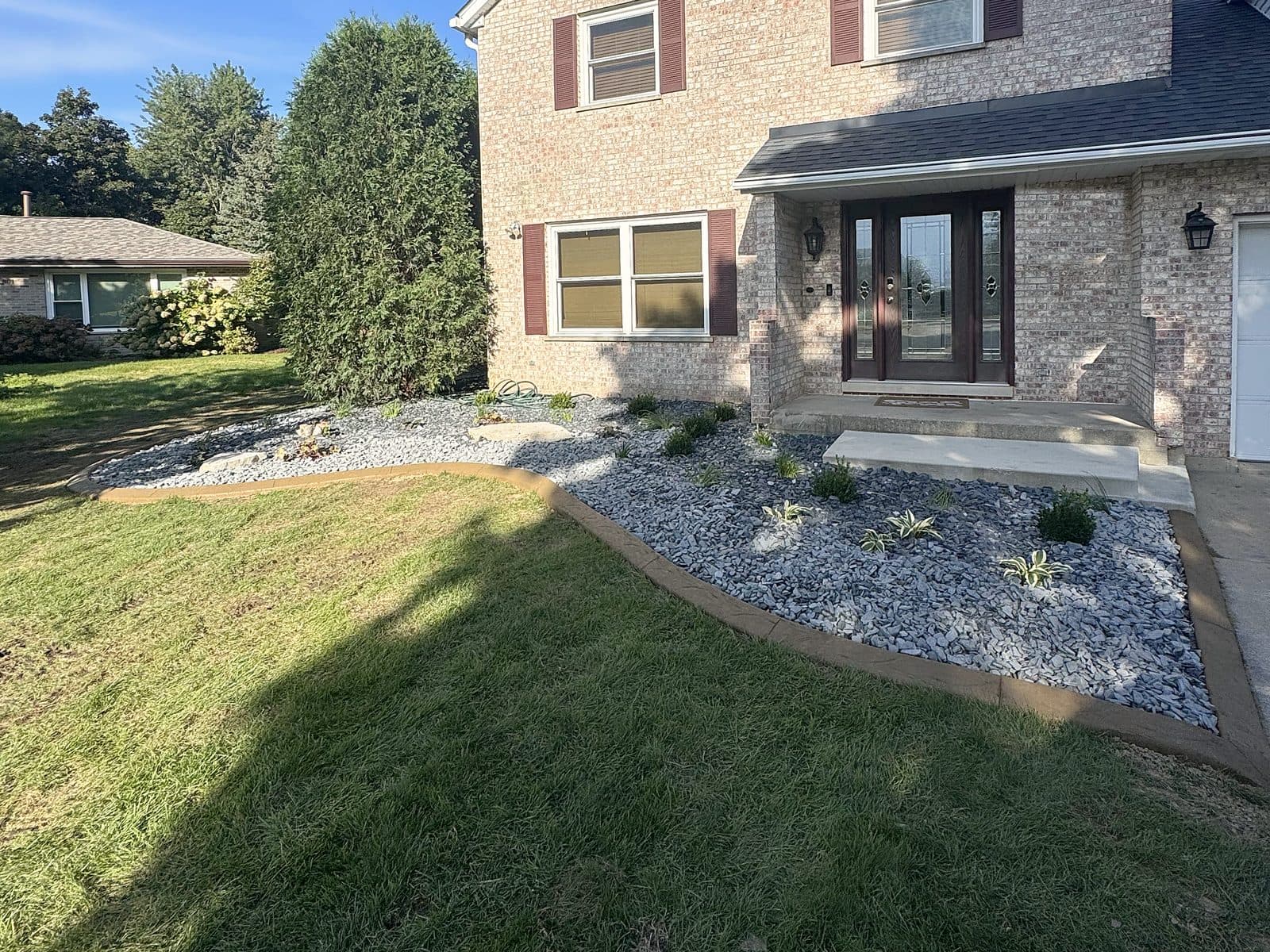 After: front yard with overgrown bushes and red rock, redone with stamped concrete curbing and blue-gray rock