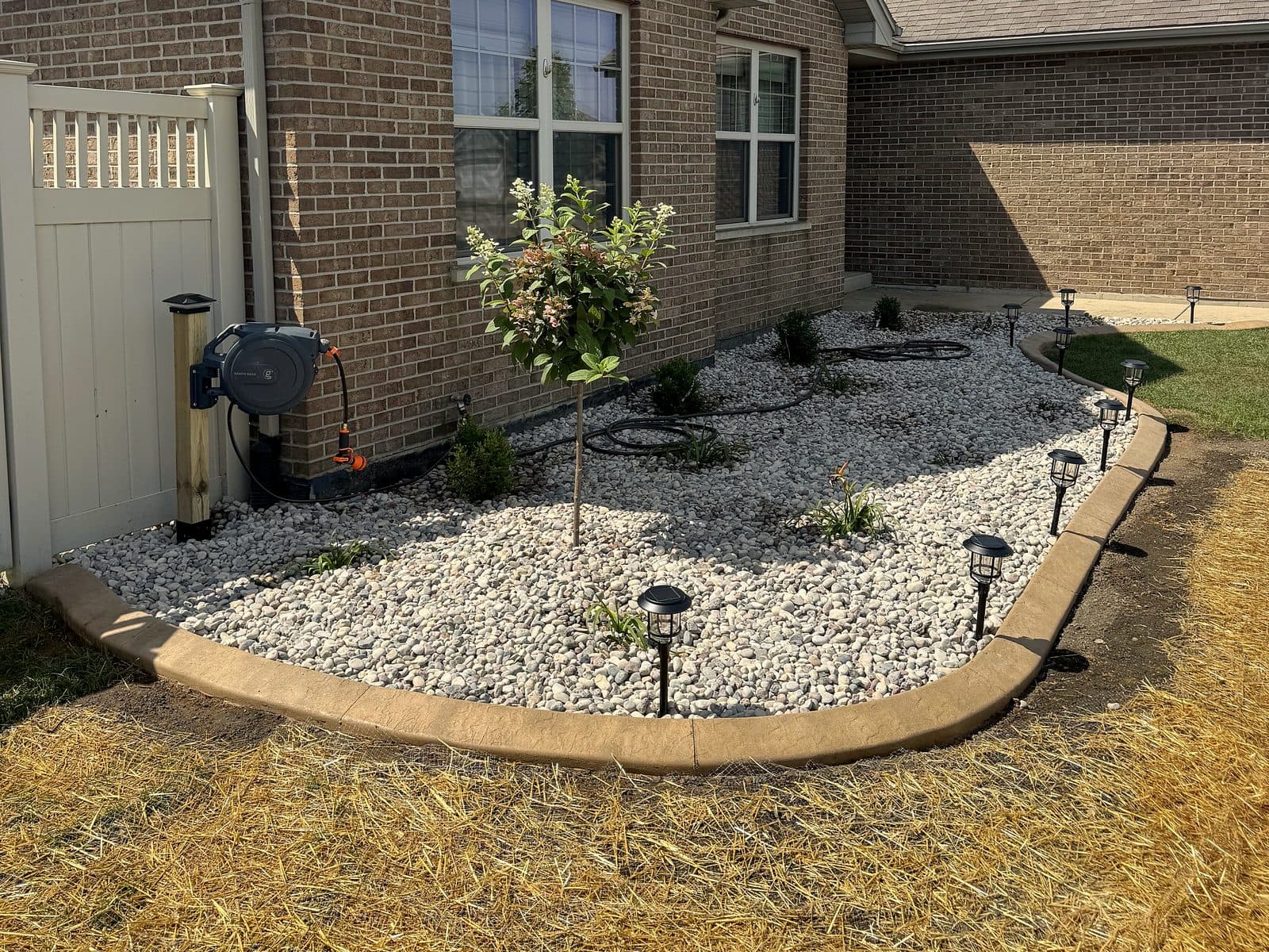 After: bare, torn-out bed along a brick house, finished with river rock, a new tree and concrete curb edging